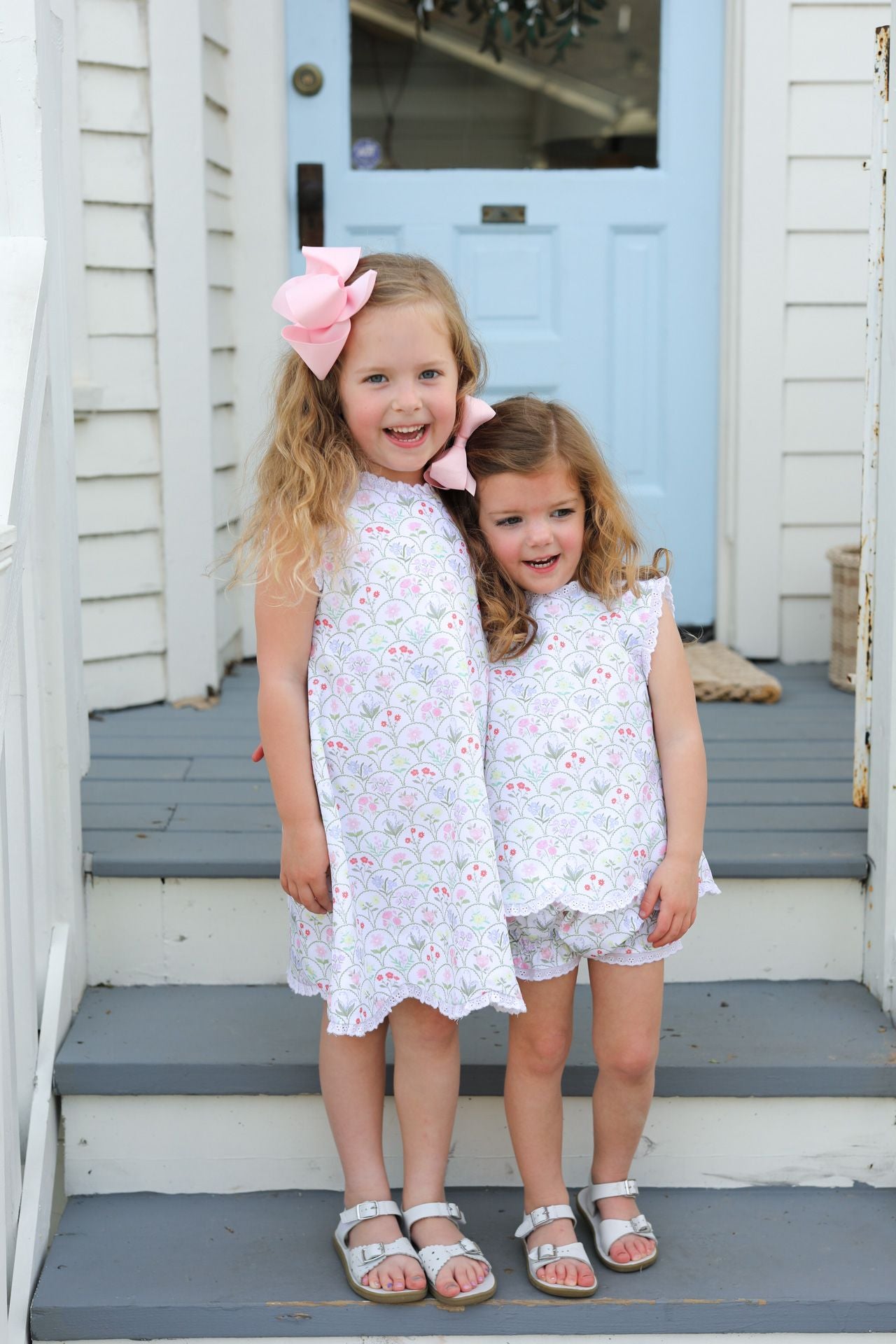 Two young girls in matching outtfits standing on a set of stairs with a blue door in the background.