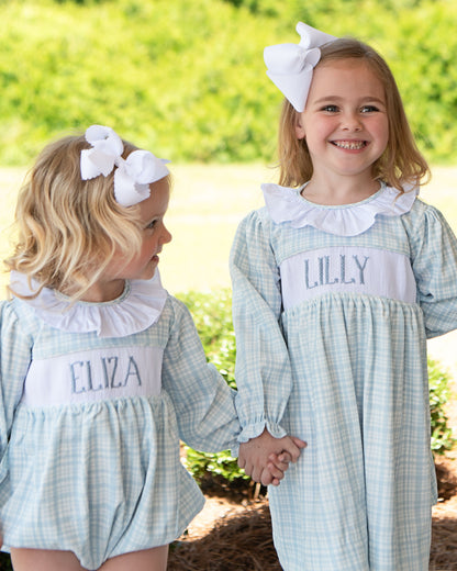 Two young girls in matching dresses with embroidered names, standing outdoors.