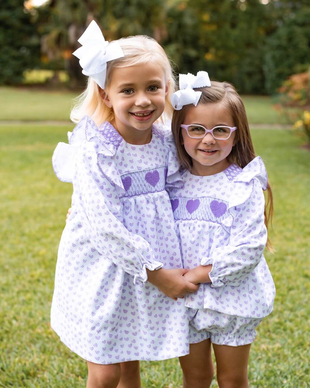 Two young girls in matching dresses with heart patterns standing outdoors.