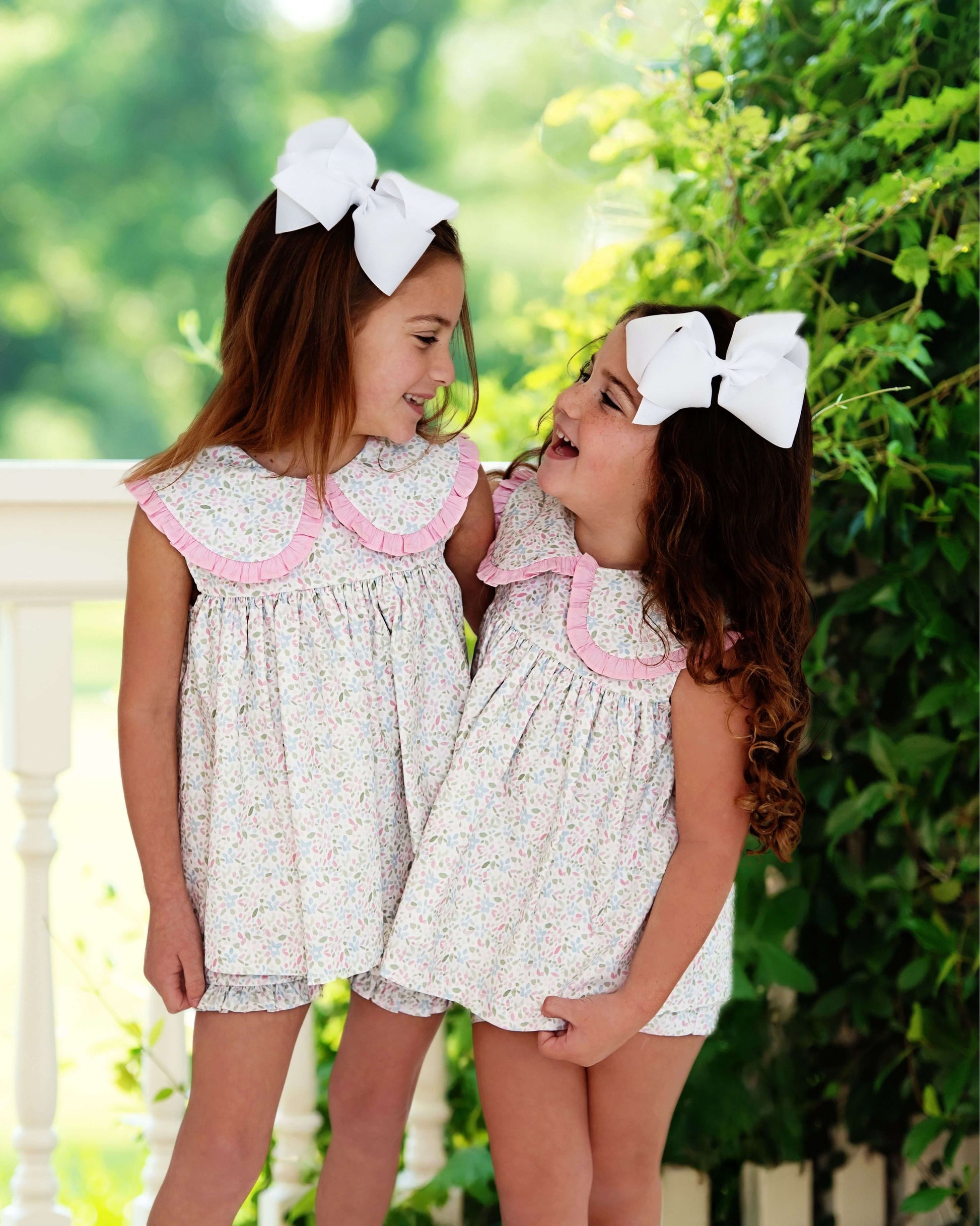 Two young girls in matching dresses with large white bows outdoors.