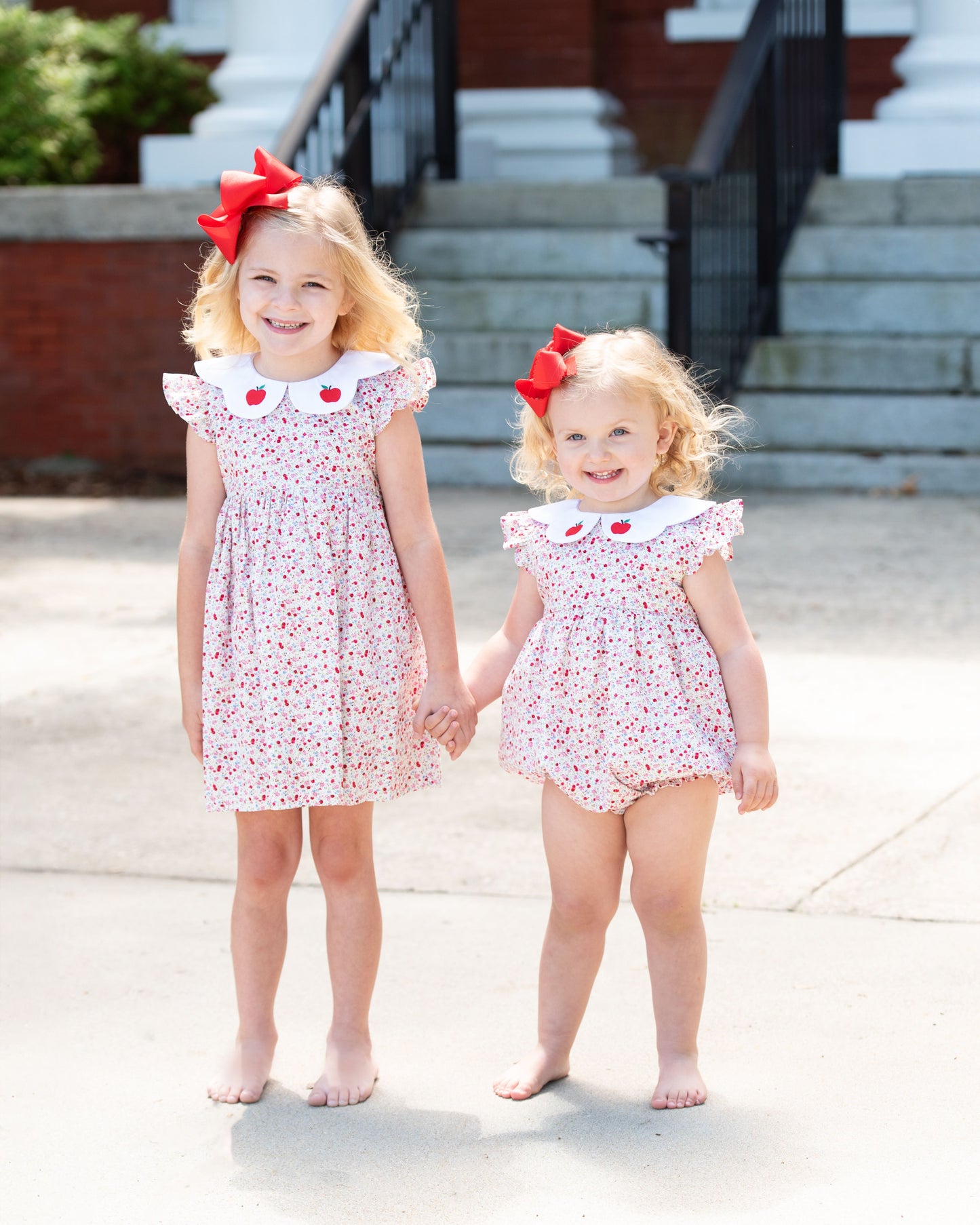 Two young girls in matching dresses with red bows standing on a sidewalk.