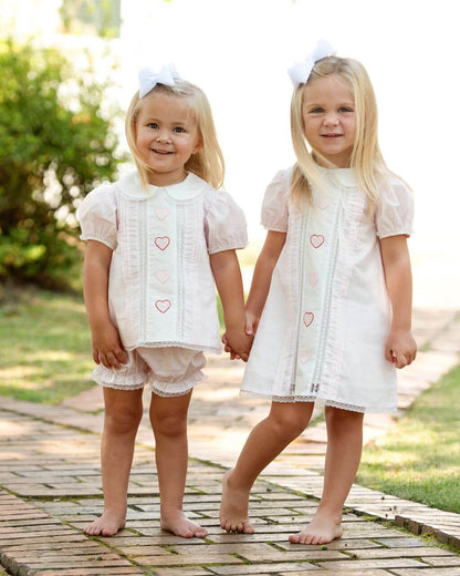 Two young girls in matching embroidered outfits with heart designs standing on a brick path outdoors.