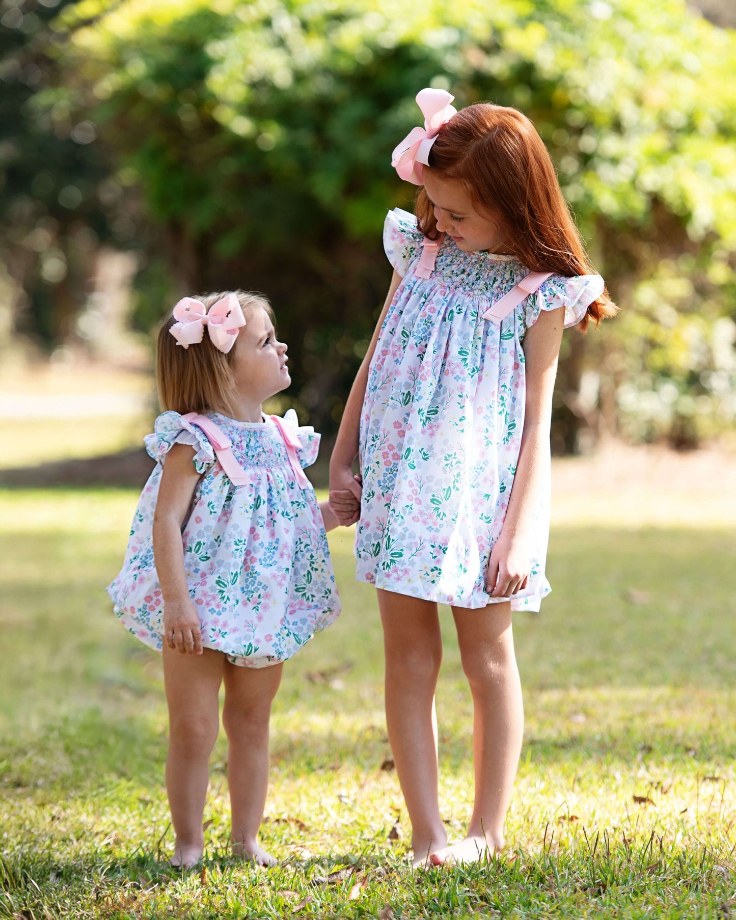 Two young girls in matching floral dresses standing outdoors on grass.