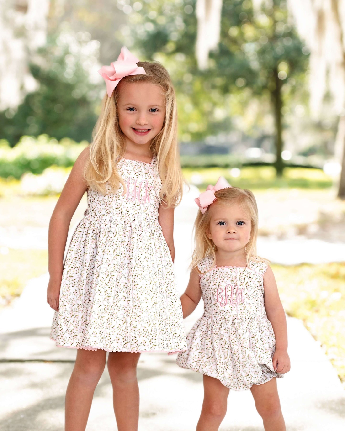 Two young girls in matching floral dresses with pink bows standing outdoors.