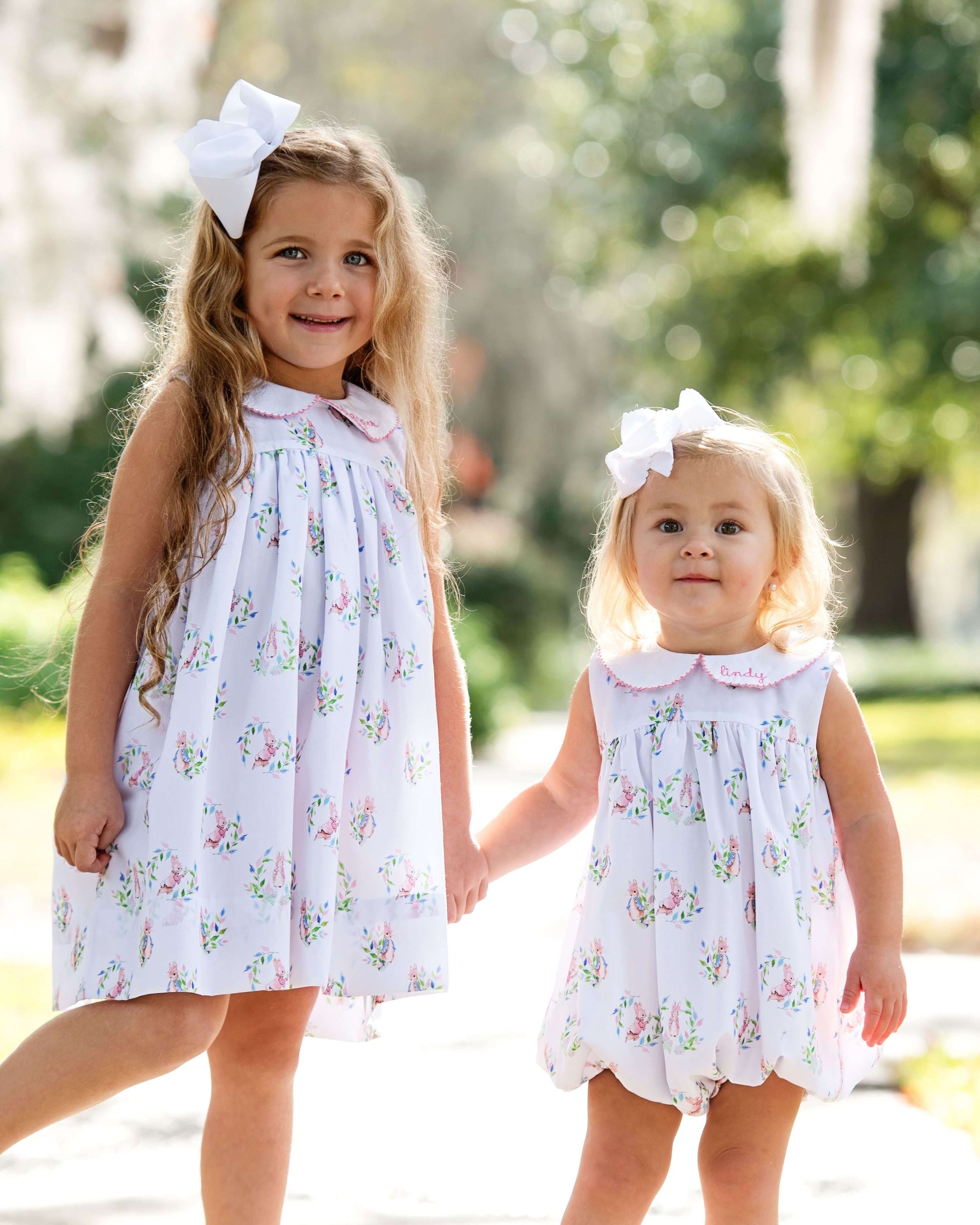 Two young girls in matching floral dresses with white bows outdoors.