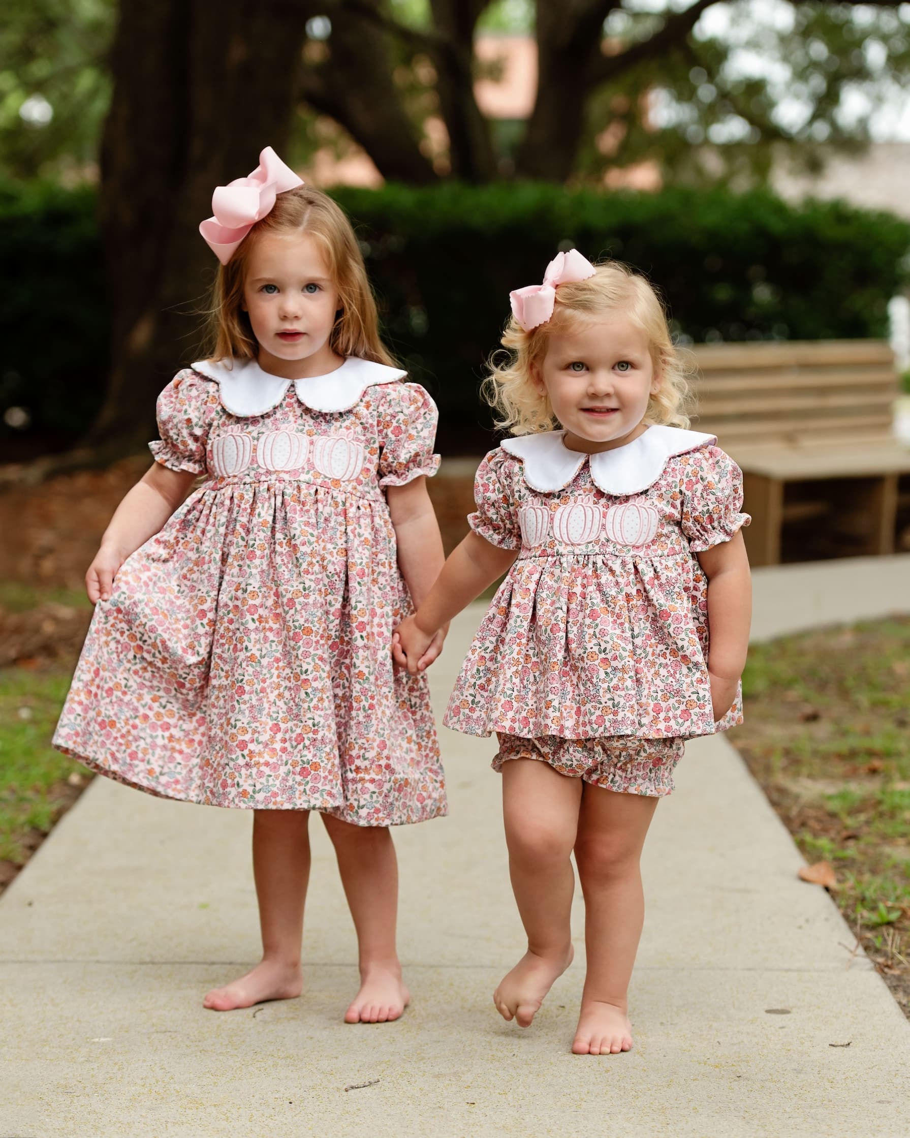 Two young girls in matching floral pumpkin outfits standing outdoors on a path.
