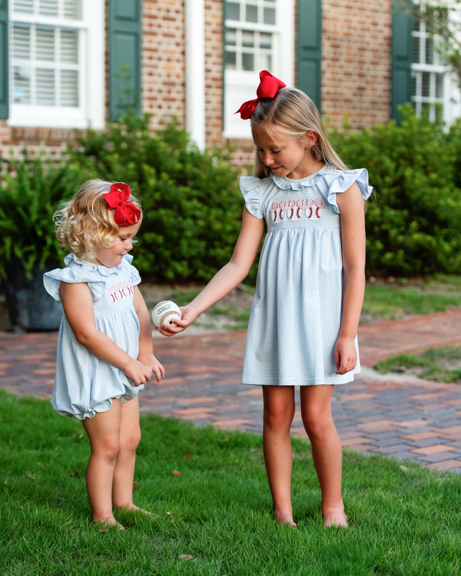 Two young girls in matching light blue dresses with red bows standing on grass.