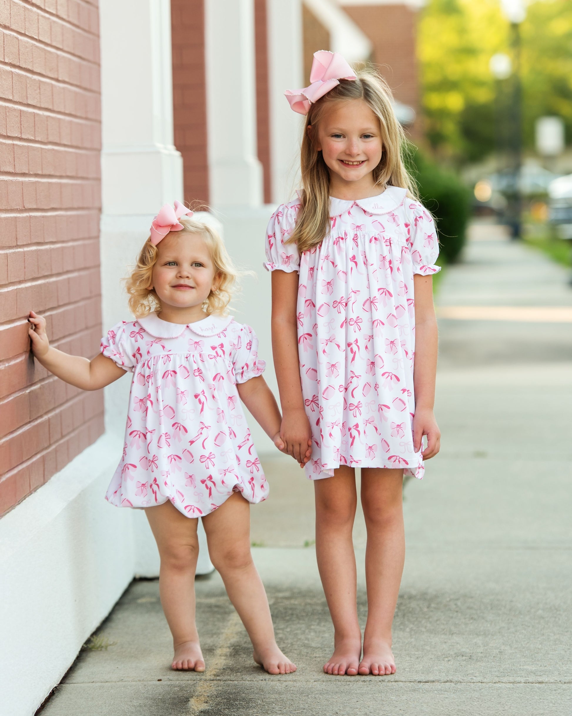 Two young girls in matching pink dresseand bubble standing on a sidewalk.
