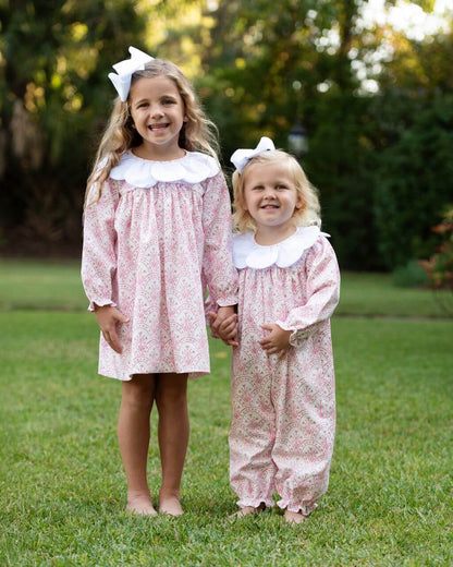 Two young girls in matching pink floral outfits with petal collars, standing on grass with trees in the background.