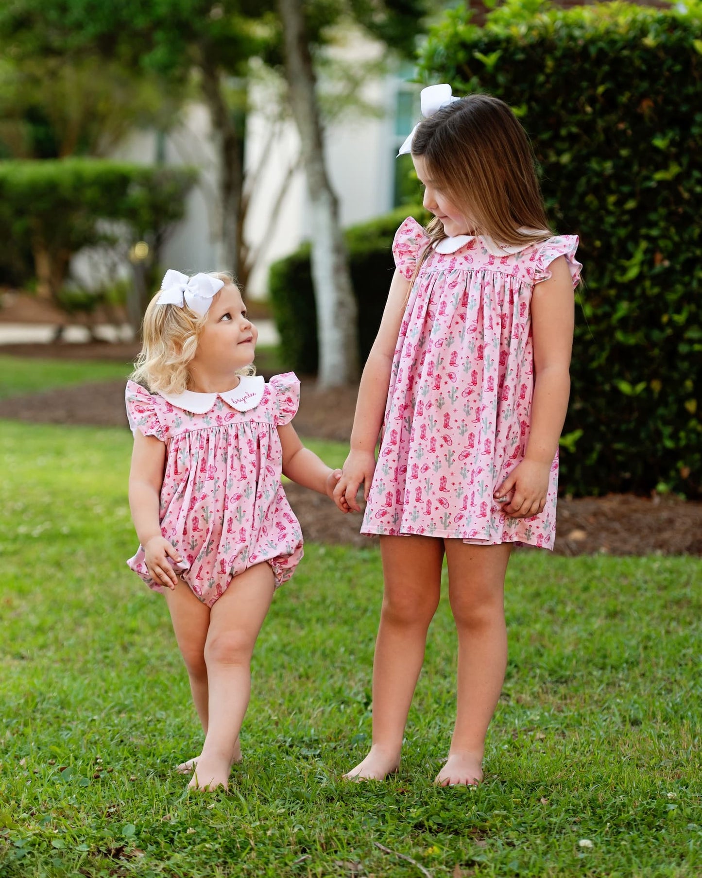 Two young girls in matching pink rodeo print dresses standing on grass.