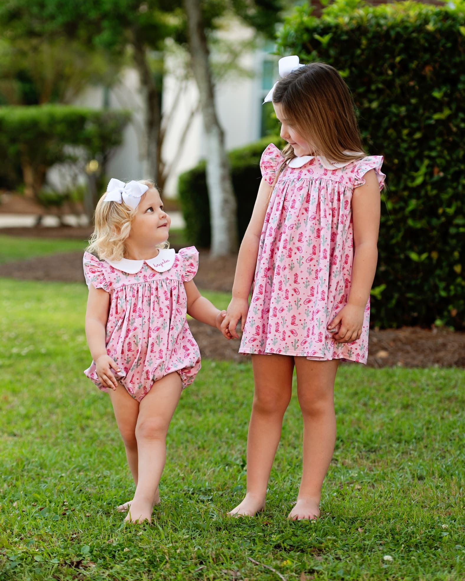 Two young girls in matching pink rodeo print dresses standing on grass.