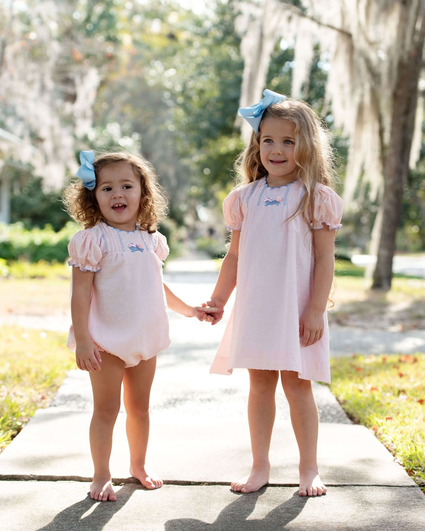 Two young girls in matching pink swiss dot outfits standing on a path outdoors.