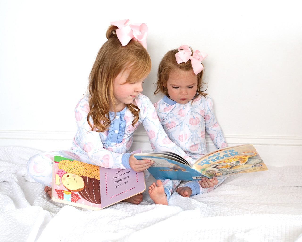 Two young girls in matching pumpkin pajama set with pink bows in their hair reading books on a white bed.