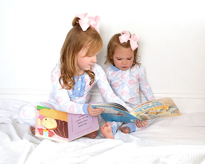 Two young girls in matching pumpkin pajama set with pink bows in their hair reading books on a white bed.