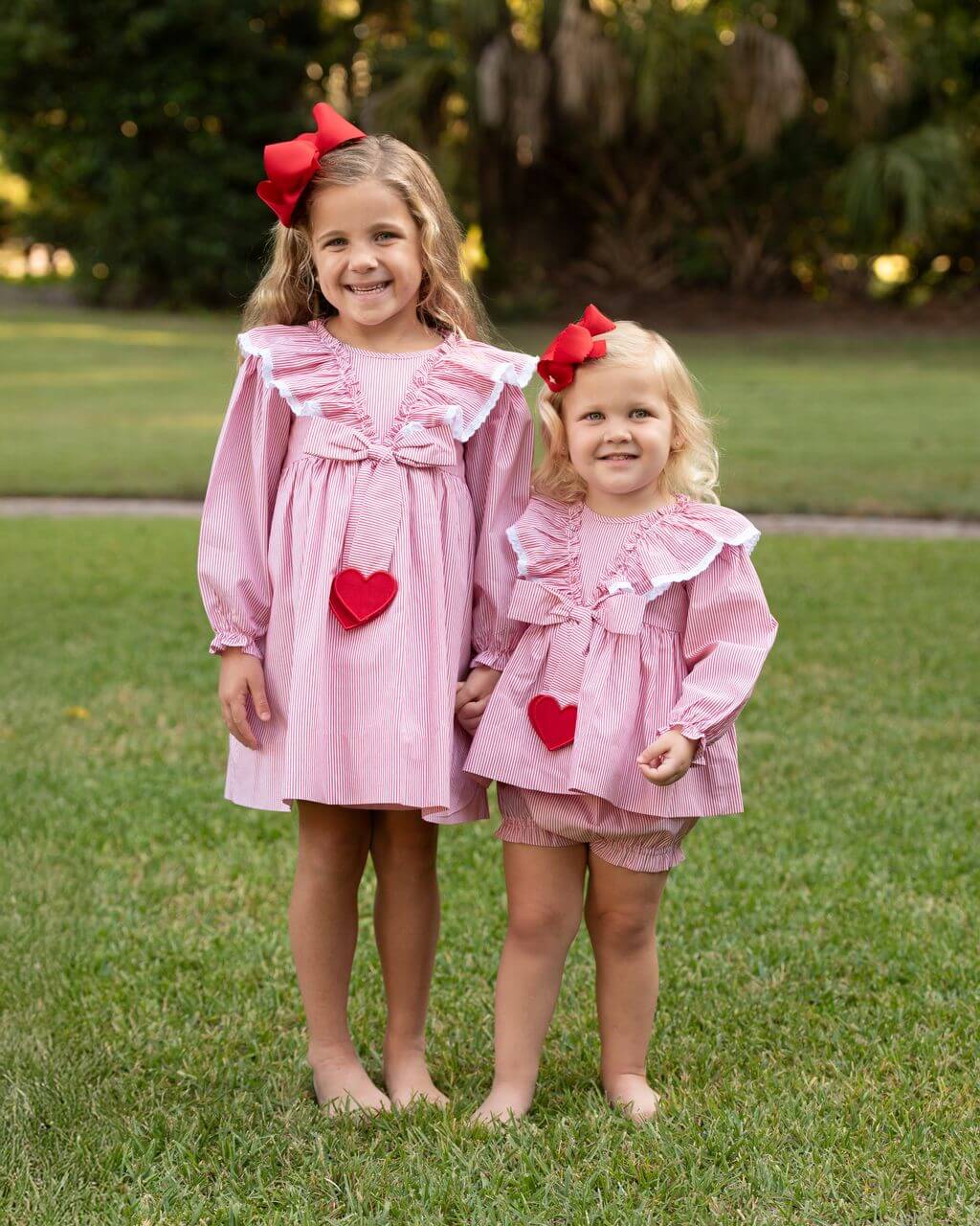 Two young girls in matching red striped dresses with red hearts standing on grass.