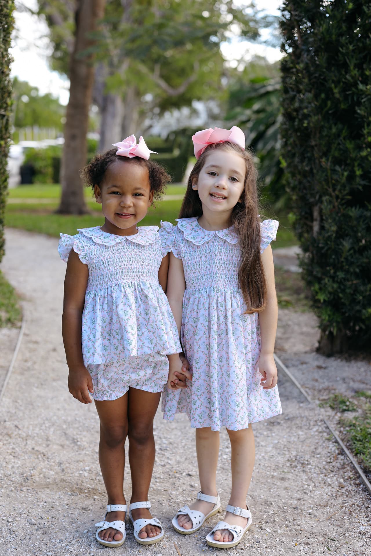 Two young girls in matching smocked outfits standing on a path with trees in the background.