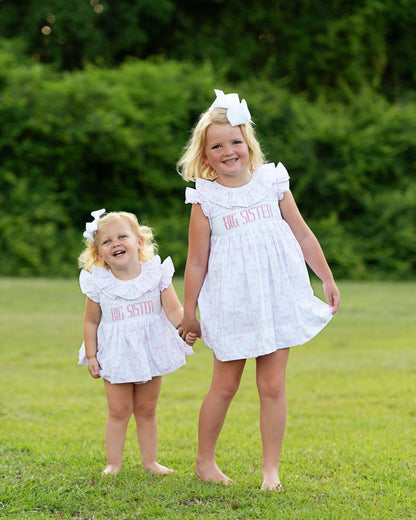 Two young girls in matching white dresses with 'Big Sister' text, standing on grass.