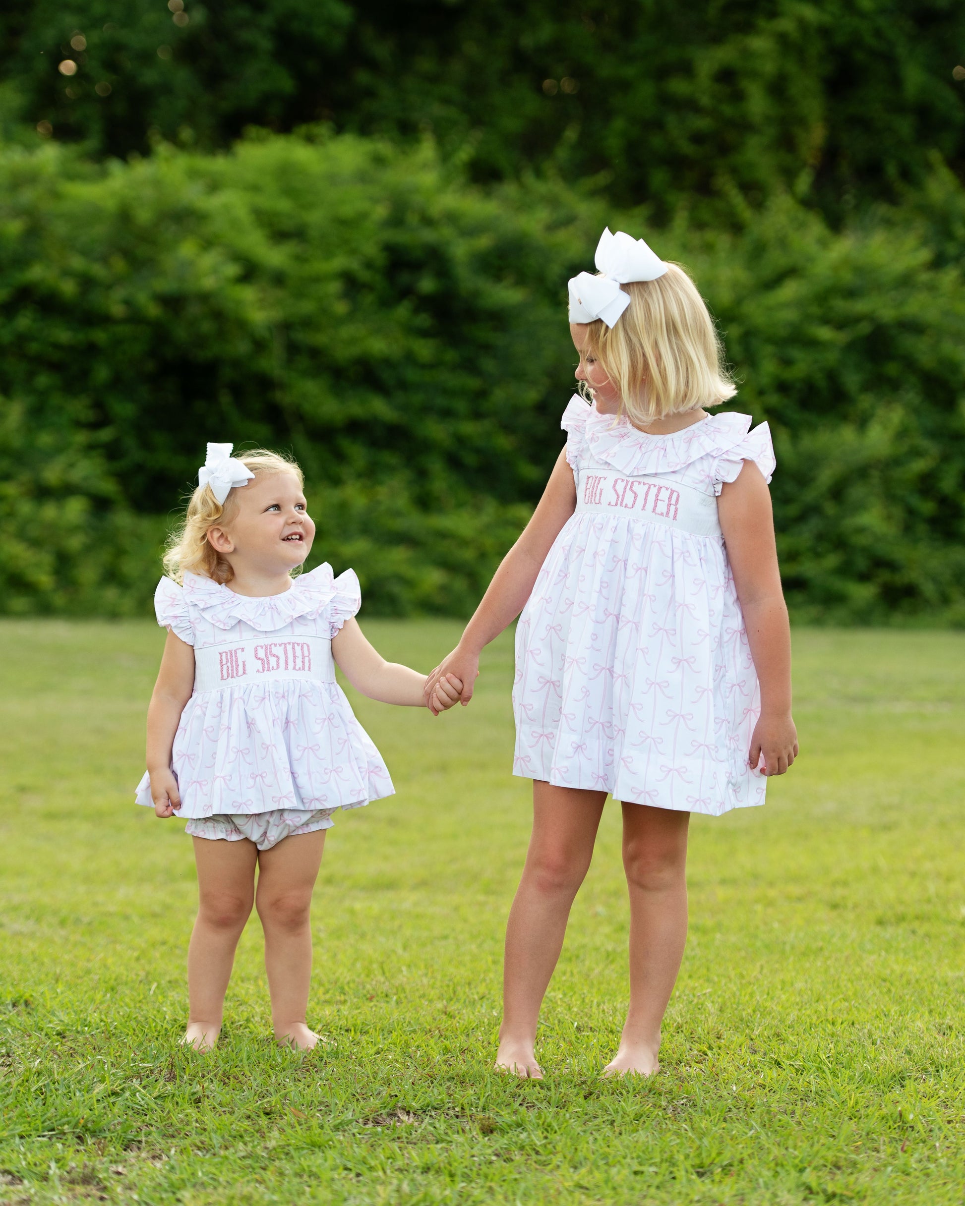 Two young girls in matching white dresses with 'Big Sister' text standing on grass.