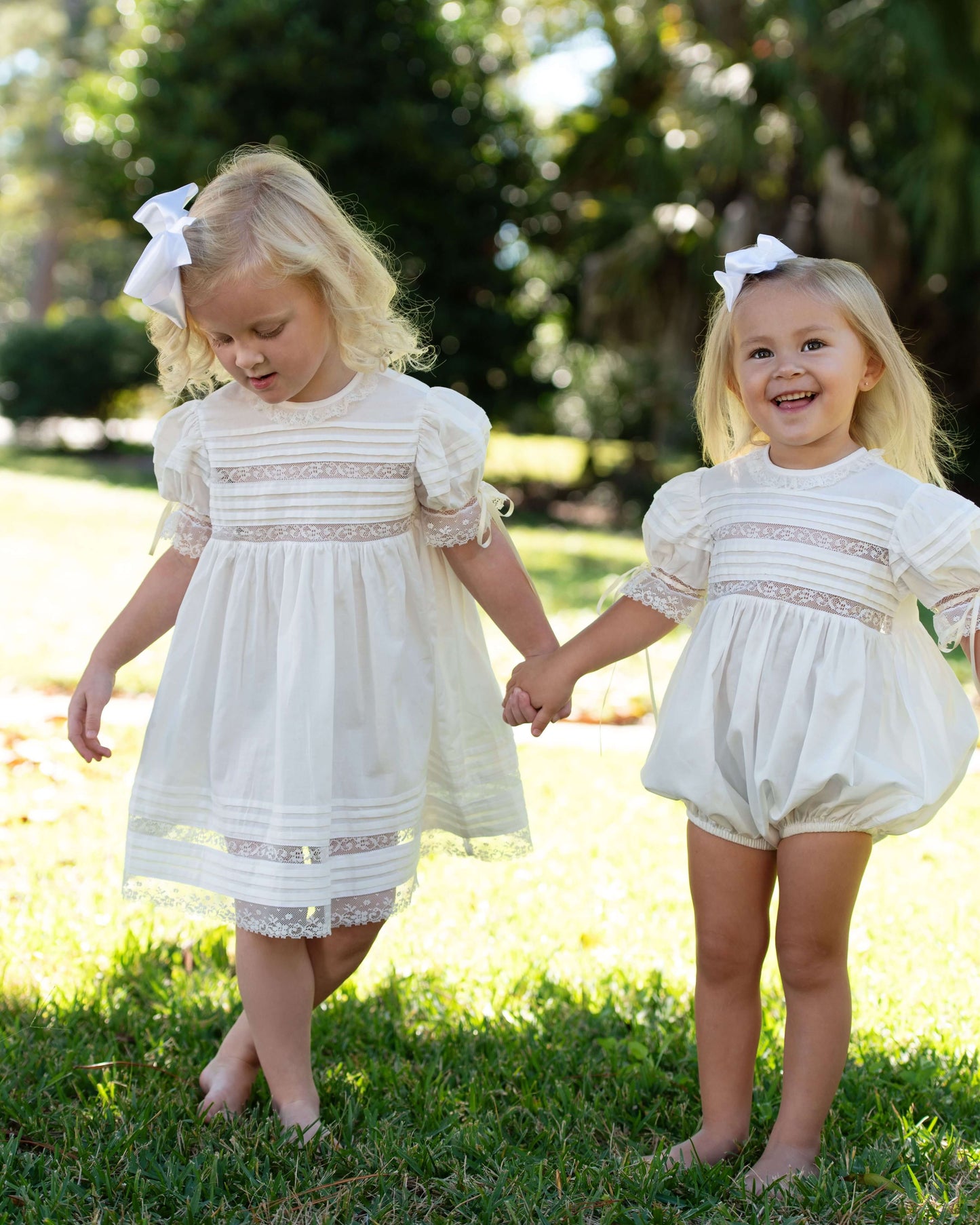 Two young girls in matching white outfits standing on grass with trees in the background.