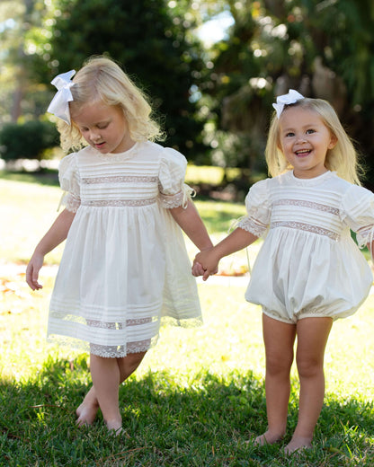 Two young girls in matching white outfits standing on grass with trees in the background.