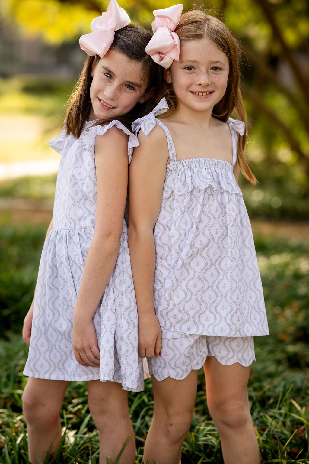 Two young girls in matching white patterned dresses standing outdoors with greenery in the background.