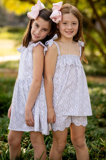 Two young girls in matching white patterned dresses standing outdoors with greenery in the background.