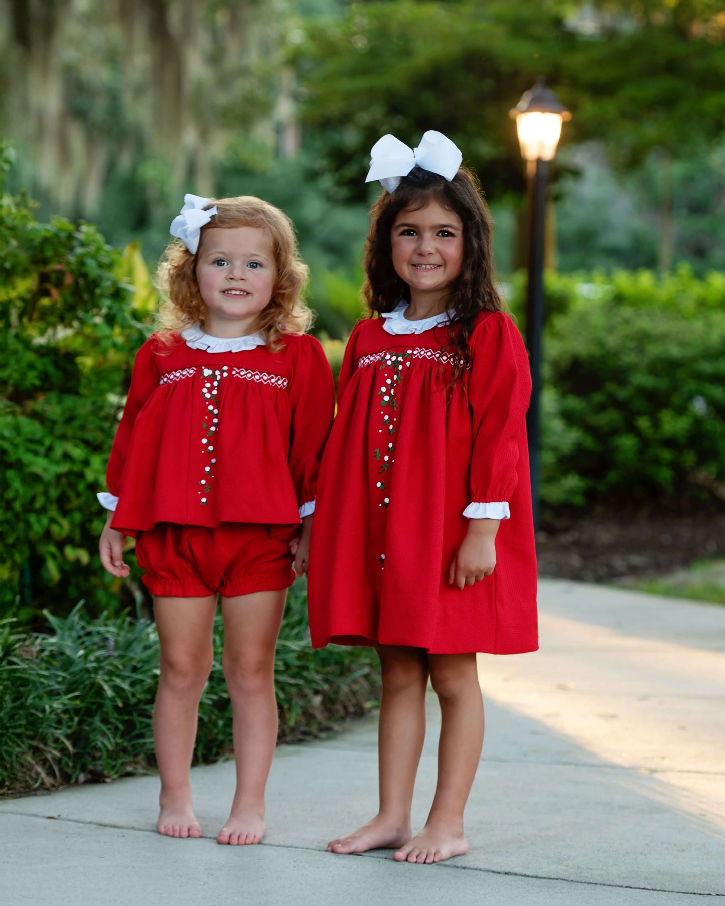 Two young girls in red outfits with white bows standing outdoors on a path.