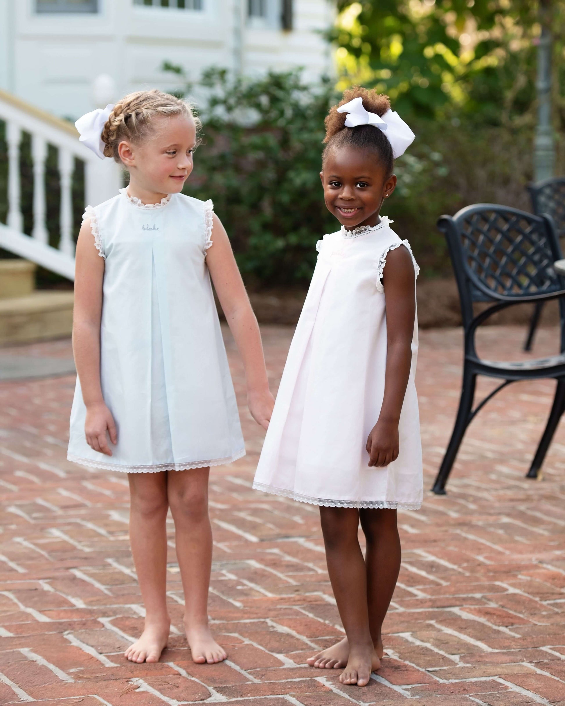 Two young girls in white dresses standing on a brick patio.