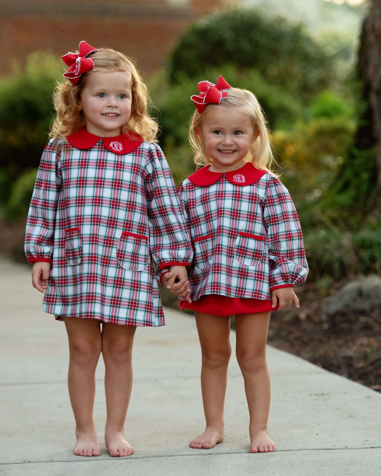 Two young girls standing barefoot on a concrete path outdoors, wearing matching long-sleeve plaid dresses with red collars and red bow hair accessories. The dresses have a white, red, and blue plaid pattern with a red embroidered monogram on the collar.