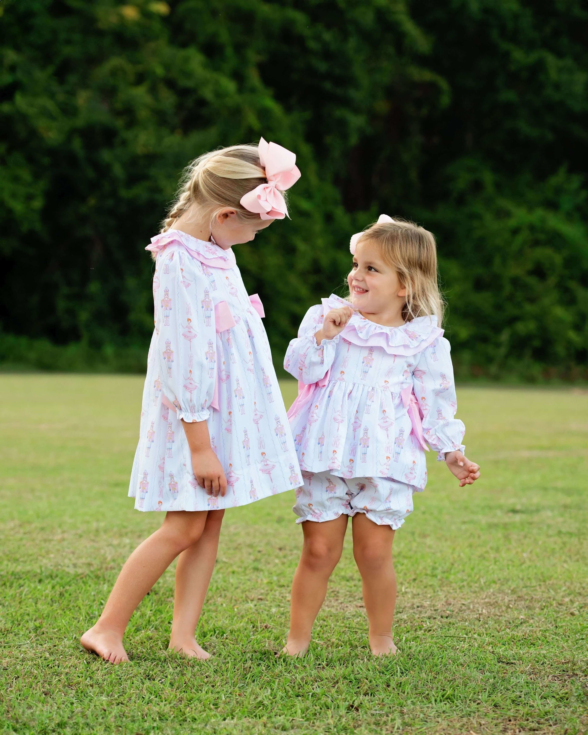Two young girls standing barefoot on a grassy lawn outdoors, wearing matching light lavender and pink floral print dresses with ruffled collars and shorts, one with a large pink bow in her hair.