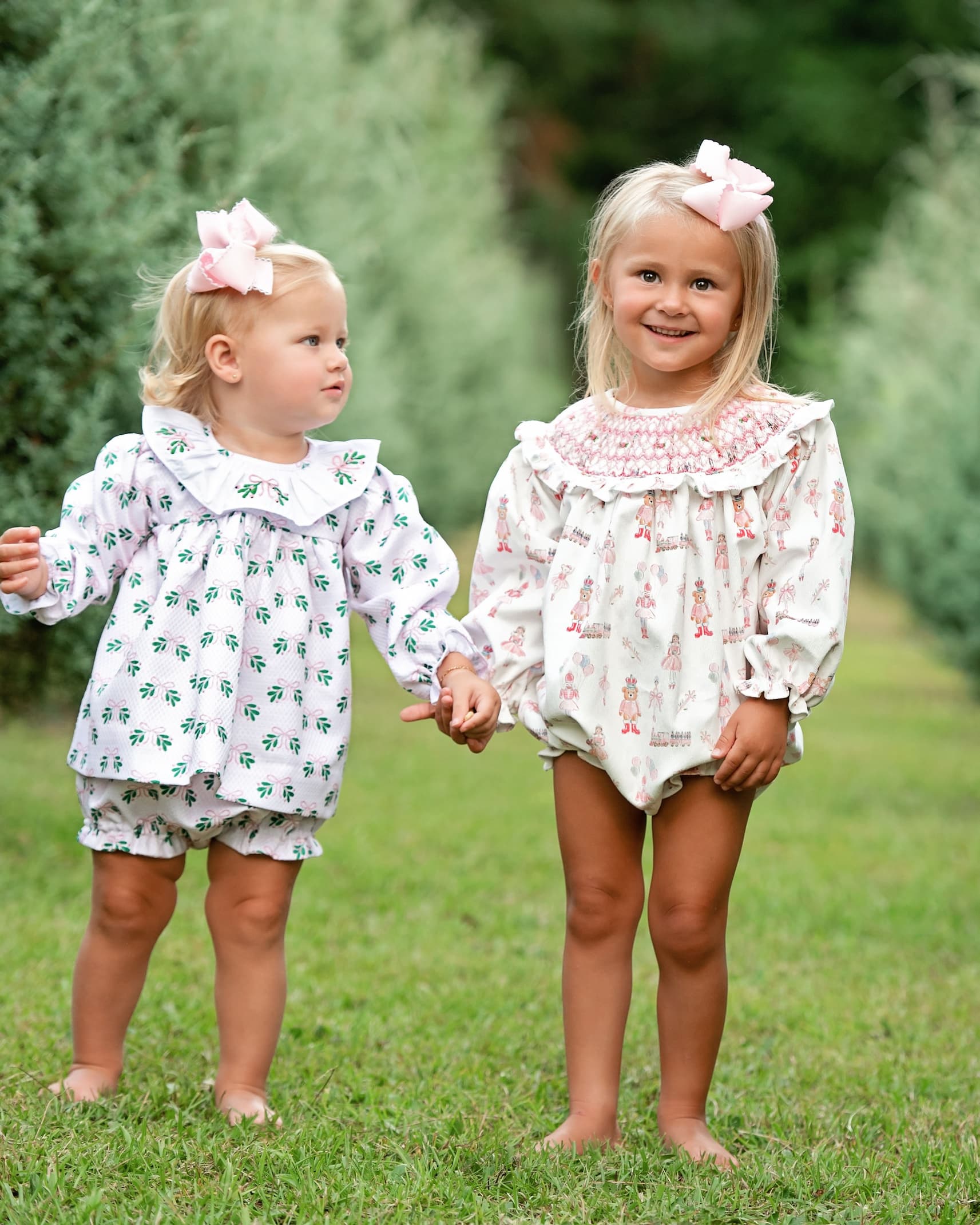 Two young girls standing barefoot on a grassy lawn outdoors, wearing matching white long-sleeve romper outfits with small pink and green floral prints and pink bows in their hair.