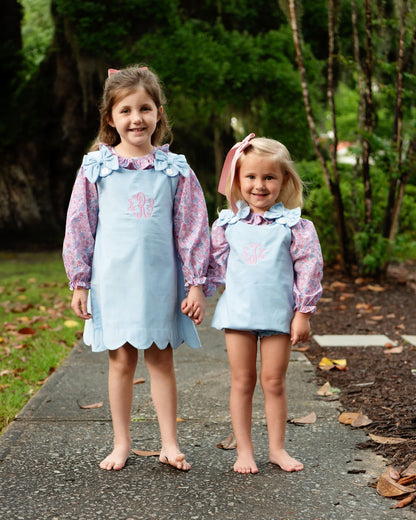 Two young girls standing barefoot on a paved path outdoors, wearing matching light blue apron-style dresses over pink floral long-sleeve shirts. The dresses have ruffled light blue necklines with blue flower appliqués and a pink monogram on the chest.