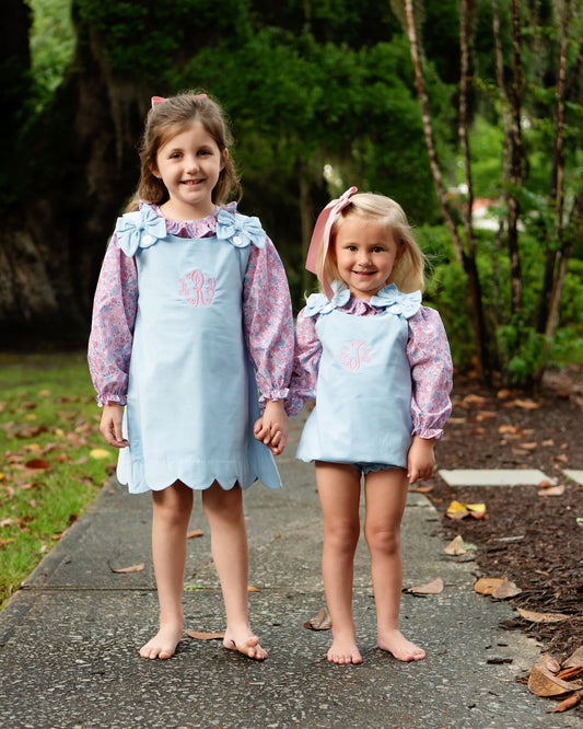 Two young girls standing barefoot on a paved path outdoors, wearing matching light blue apron-style dresses over pink floral long-sleeve shirts. The dresses have ruffled light blue necklines with blue flower appliqués and a pink monogram on the chest.