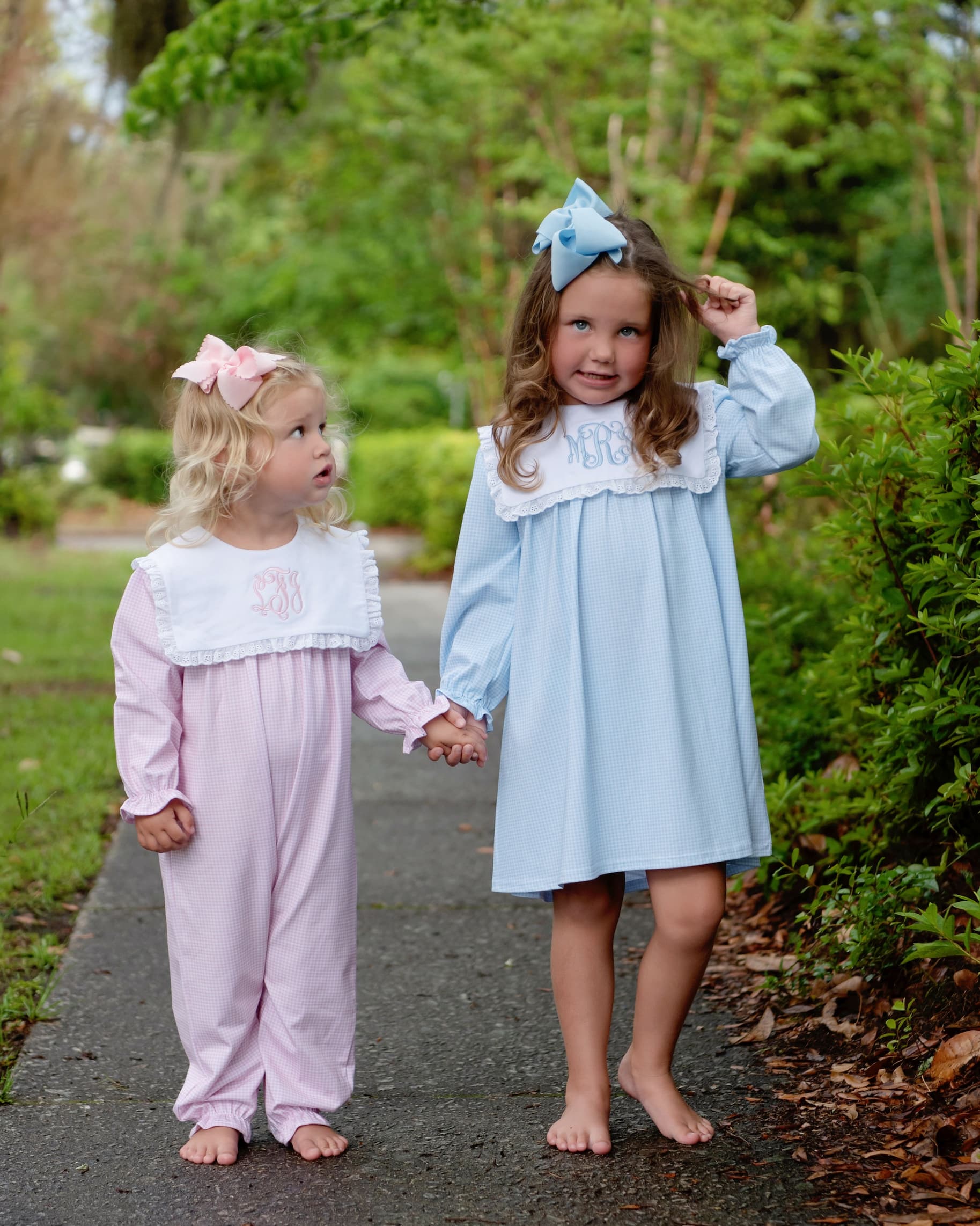 Two young girls standing barefoot on a paved path outdoors, wearing matching long-sleeve sleepwear sets. The girl on the right wears a light blue nightgown with a white ruffled collar and monogrammed lettering, and the girl on the left wears a pale pink footed sleeper with a white ruffled collar and monogrammed lettering.