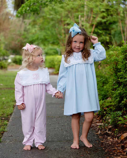 Two young girls standing barefoot on a paved path outdoors, wearing matching long-sleeve sleepwear sets. The girl on the right wears a light blue nightgown with a white ruffled collar and monogrammed lettering, and the girl on the left wears a pale pink footed sleeper with a white ruffled collar and monogrammed lettering.