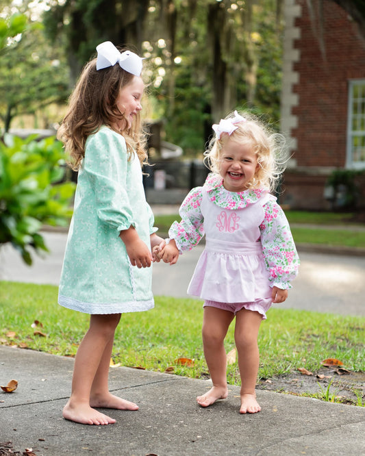 Two young girls standing barefoot on a sidewalk outdoors, holding hands. The girl on the left wears a mint green long-sleeve dress with a white bow in her hair.