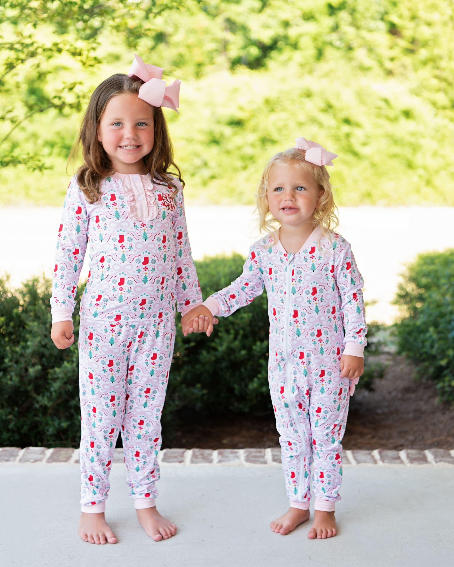 Two young girls standing barefoot outdoors and holding hands, wearing matching long-sleeve footless pajamas with a white base and a red and green floral print, each with a pink bow in their hair.
