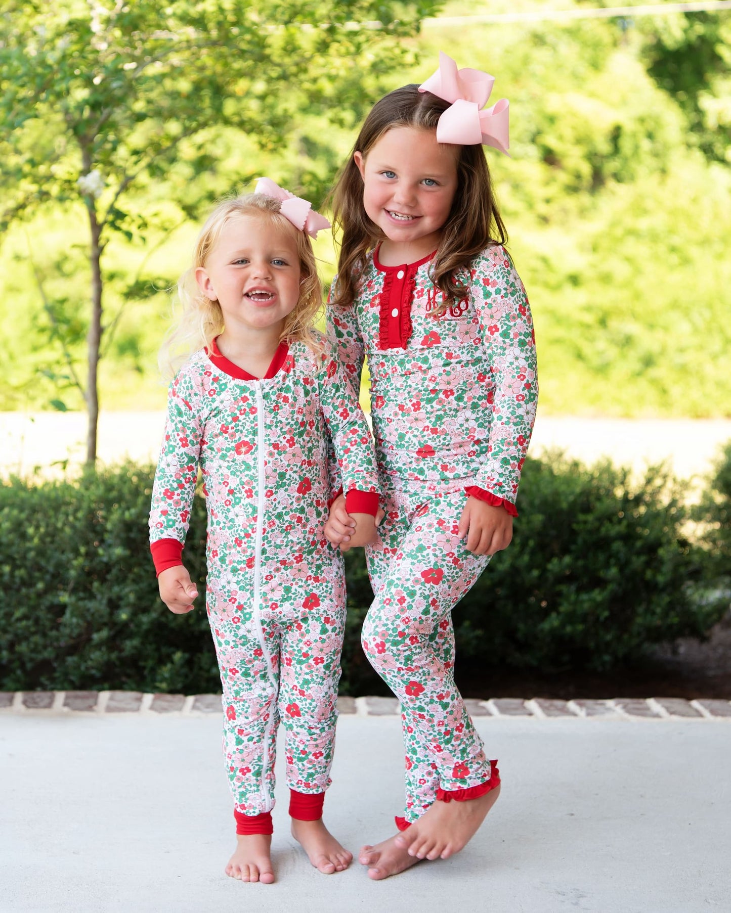 Two young girls standing barefoot outdoors, wearing matching floral one-piece pajamas with red trim and pink bows in their hair. The pajamas have a white base with a red and green floral pattern.