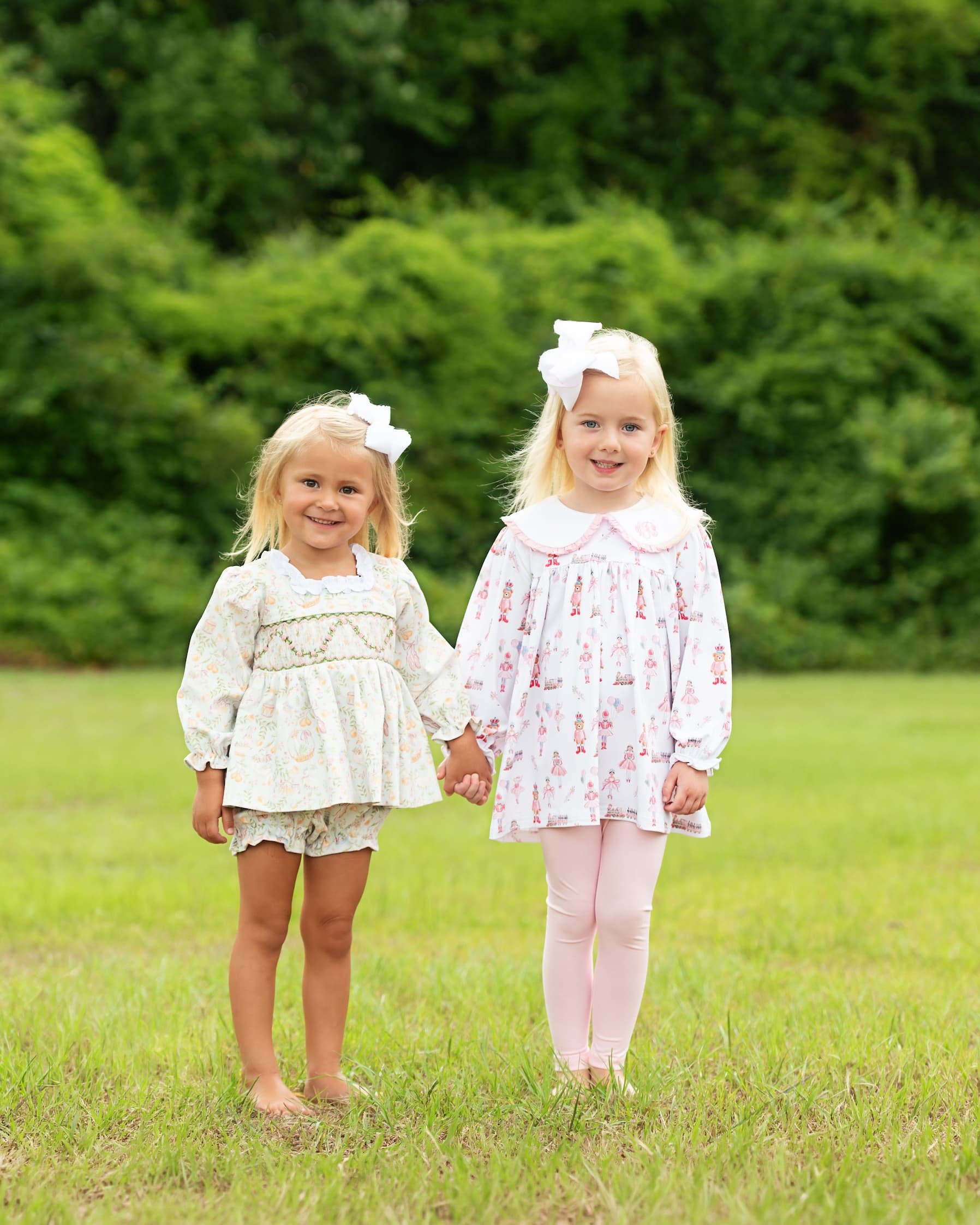 Two young girls standing in a grassy field wearing matching dresses with pink accents and white bows in their hair.