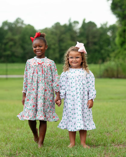 Two young girls standing on a grassy field outdoors, wearing matching long-sleeve floral dresses. The girl on the left has dark skin and a red bow in her hair, wearing a light green dress with pink and red floral patterns.