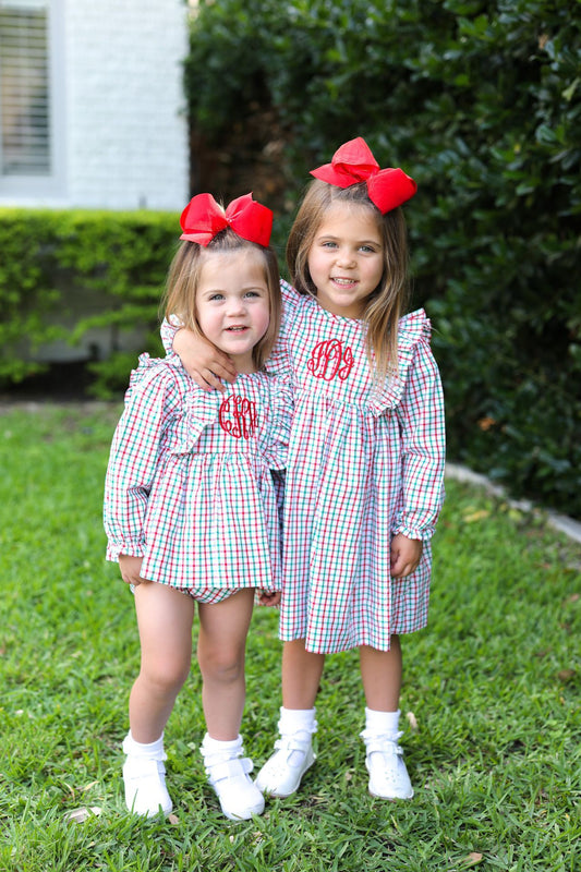 Two young girls standing on a green lawn wearing matching outfits with red monogrammed bibs and large red bows in their hair.