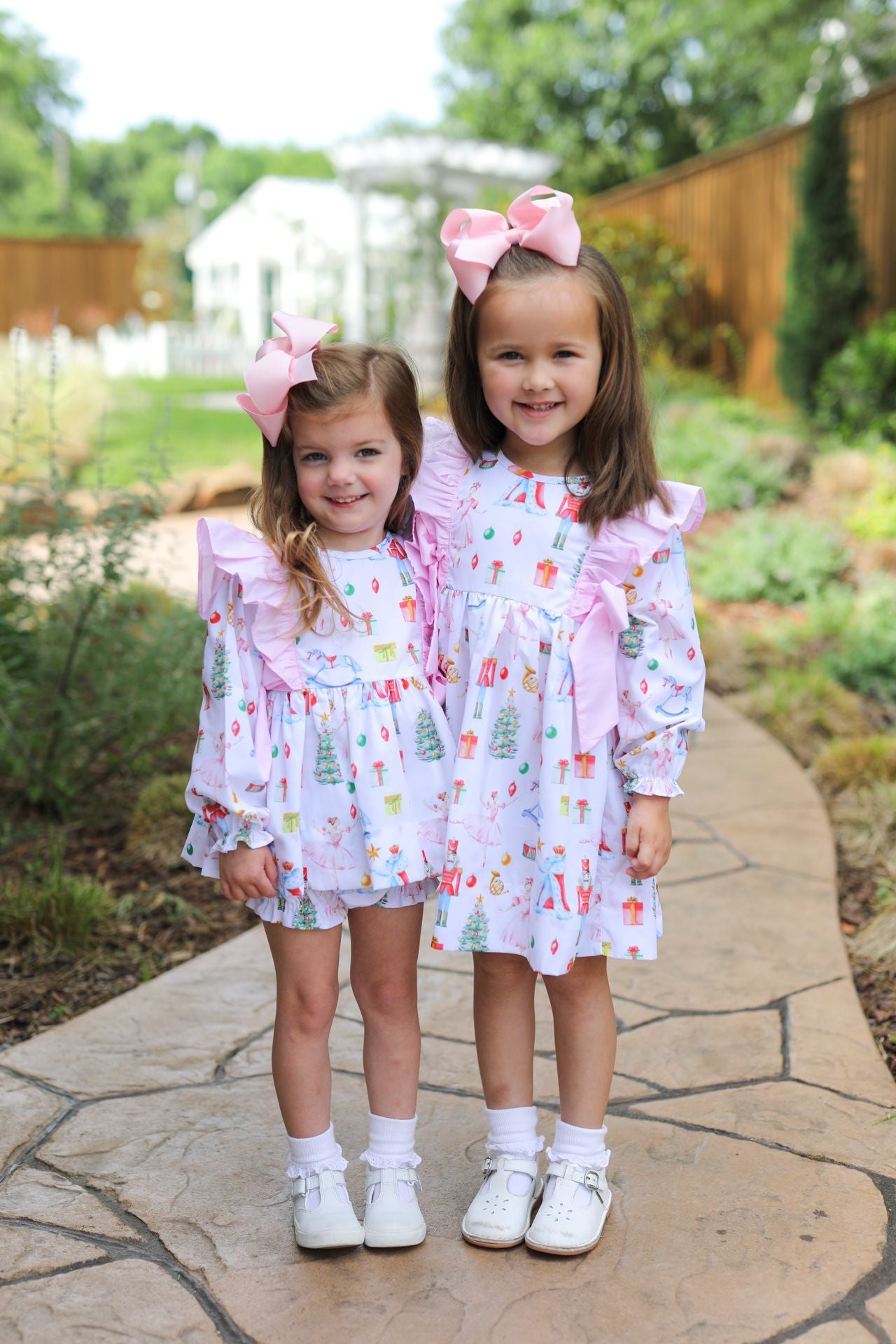 Two young girls standing on a stone path in a garden, wearing matching white dresses with a colorful Christmas tree print and pink ruffled trim, along with pink bows in their hair and white shoes.