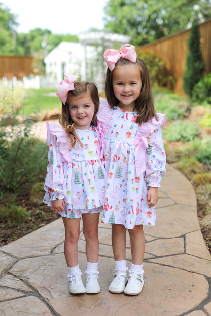 Two young girls standing on a stone path in a garden, wearing matching white dresses with a colorful Christmas tree print and pink ruffled trim, along with pink bows in their hair and white shoes.