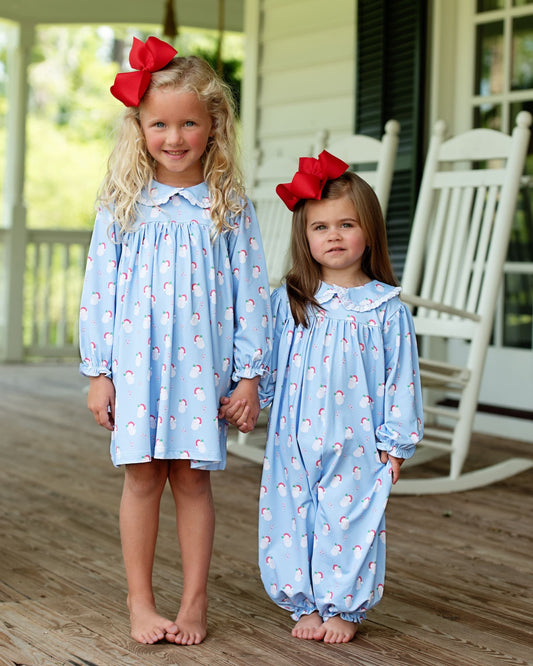Two young girls standing on a wooden porch holding hands, wearing matching light blue long-sleeve sleep dresses with a small red and white snowman print. The girl on the left wears a knee-length dress and a large red bow in her hair, while the girl on the right wears a full-length footless jumpsuit and a similar red bow.