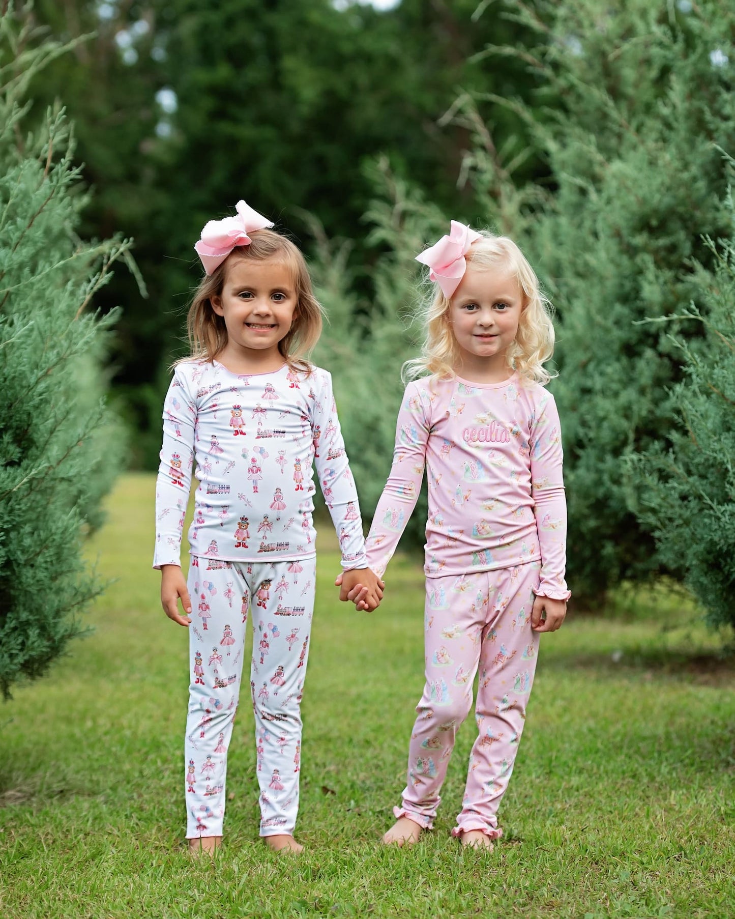 Two young girls standing outdoors in a grassy area between green trees, holding hands and wearing matching long-sleeve pajamas. The girl on the left wears a white pajama set with a pink bunny print and a large pink bow in her hair.