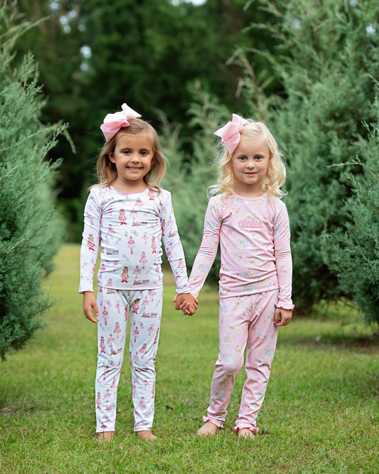 Two young girls standing outdoors in a grassy area between green trees, holding hands and wearing matching long-sleeve pajamas. The girl on the left wears a white pajama set with a pink bunny print and a large pink bow in her hair.