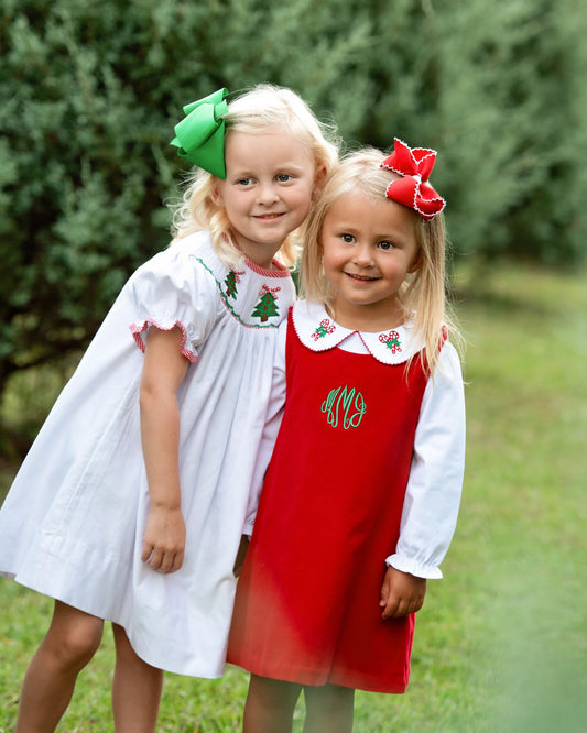 Two young girls standing outdoors in front of green trees, wearing white long-sleeve dresses with red and green holiday embroidery. One dress has a green Christmas tree motif and a green monogram, and the other has a red monogram.