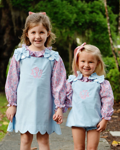 Two young girls standing outdoors in matching light blue apron-style dresses with pink floral long sleeves and bow details, each with a pink monogram on the chest.