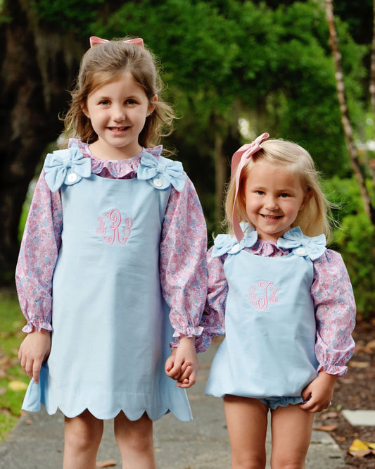 Two young girls standing outdoors in matching light blue apron-style dresses with pink floral long sleeves and bow details, each with a pink monogram on the chest.