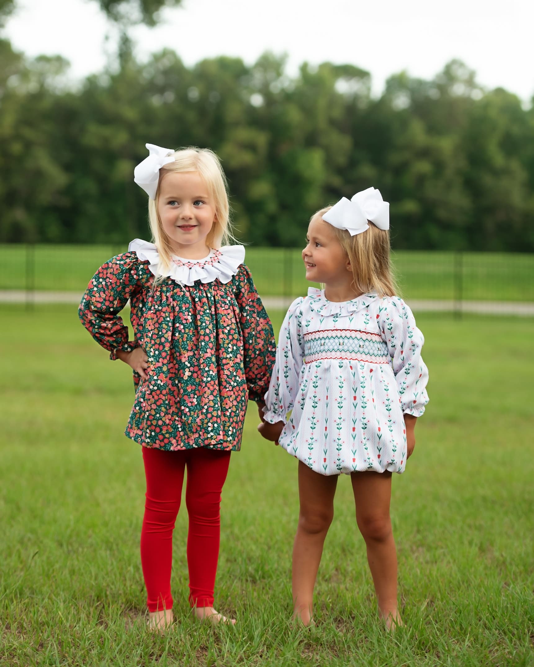 Two young girls standing outdoors on a grassy field, each wearing a long-sleeve floral or patterned dress with a ruffled collar and a large white bow in their hair. The girl on the left wears red leggings, and the girl on the right is barefoot.